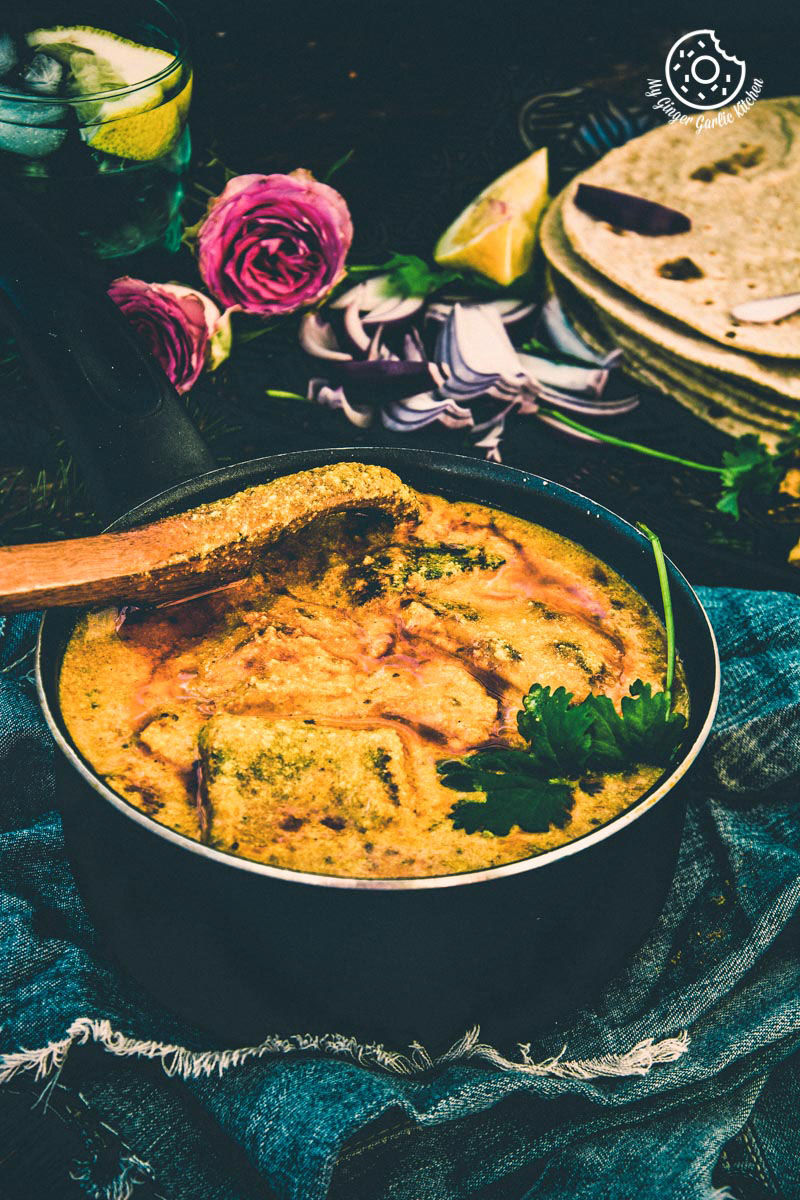 a pan of besan ke cheele ki sabzi on a table with a wooden spoon and some roses in background
