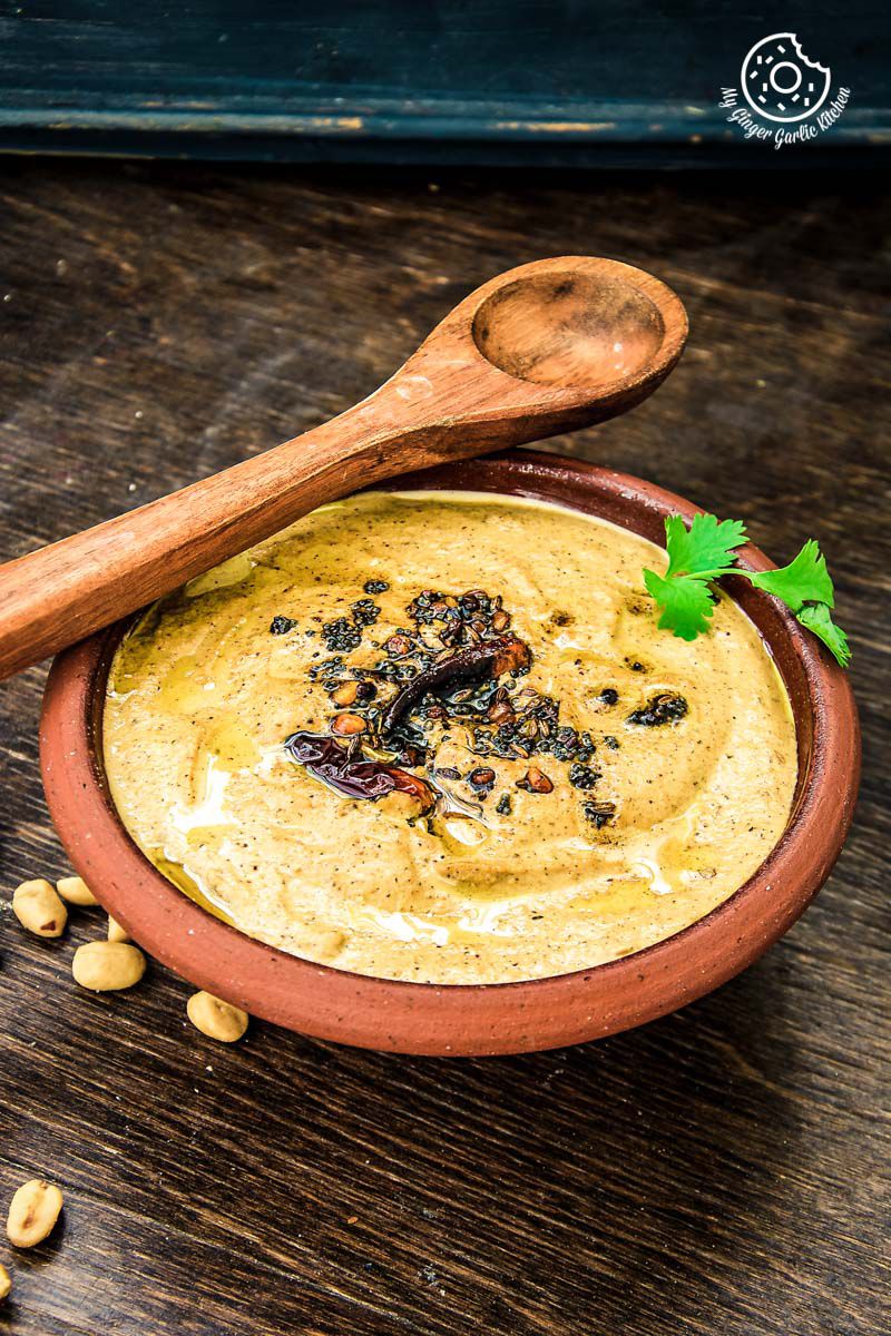 a close up of a bowl of south indian style peanut garlic chutney with a green garnish, a spoon and some peanuts