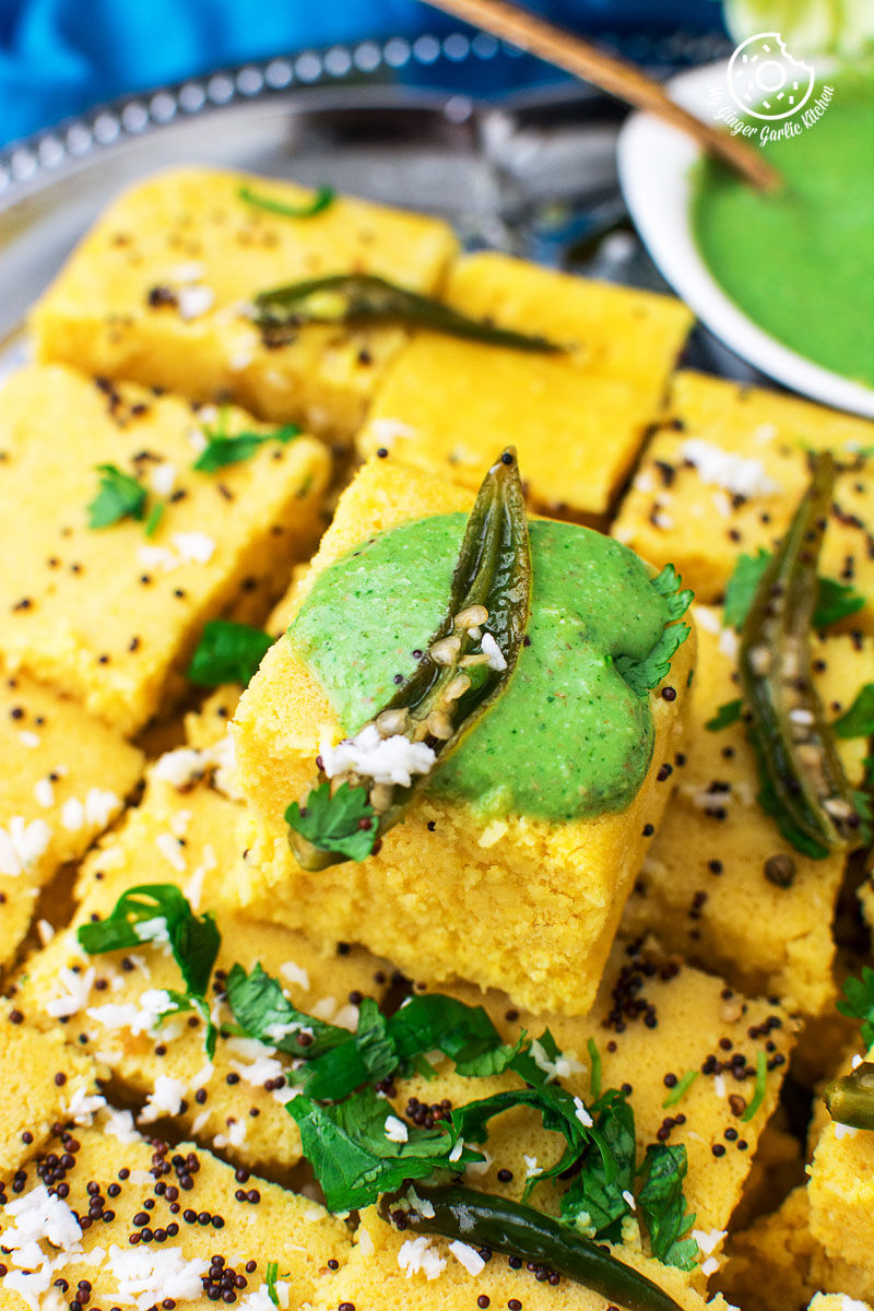 a plate of khaman dhokla with a bowl of green cilantro chutney with a green chutney on it