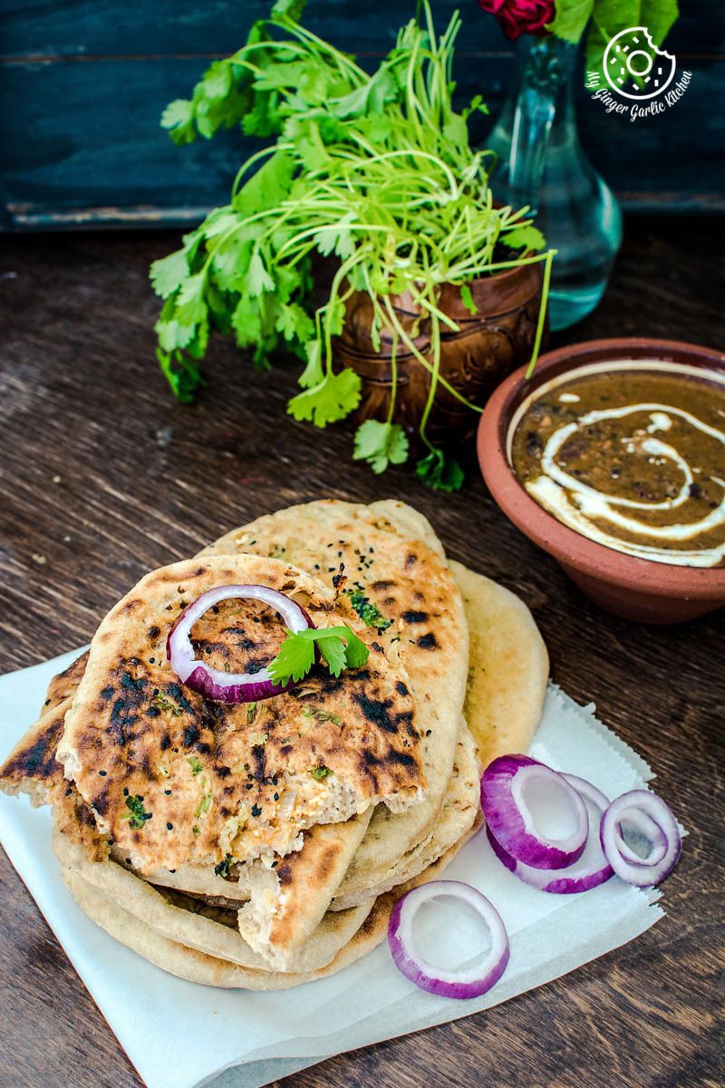 a stack of paneer stuffed naan with onions, a bowl of curry, and some cilantro leaves in background