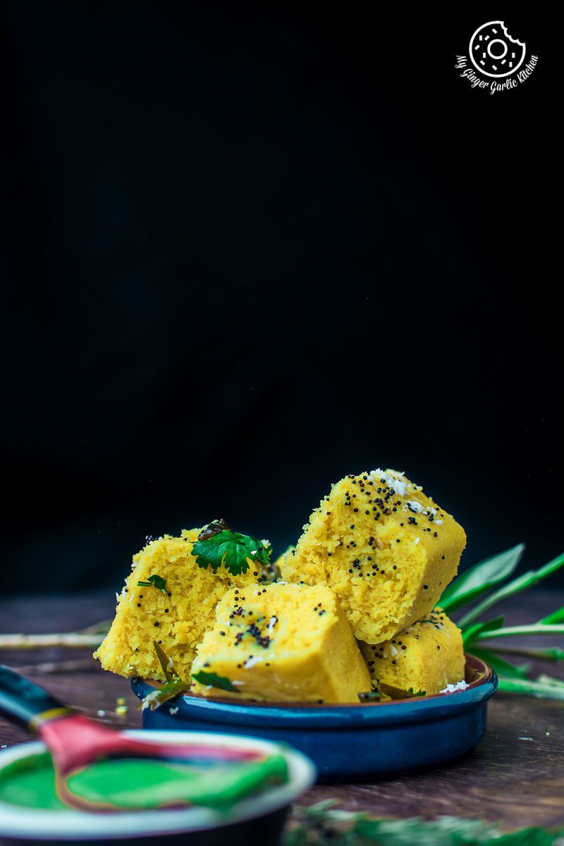 a bowl of soft and spongy khaman dhokla topped with tempering and fresh cilantro with a spoon and a bowl of green chutney