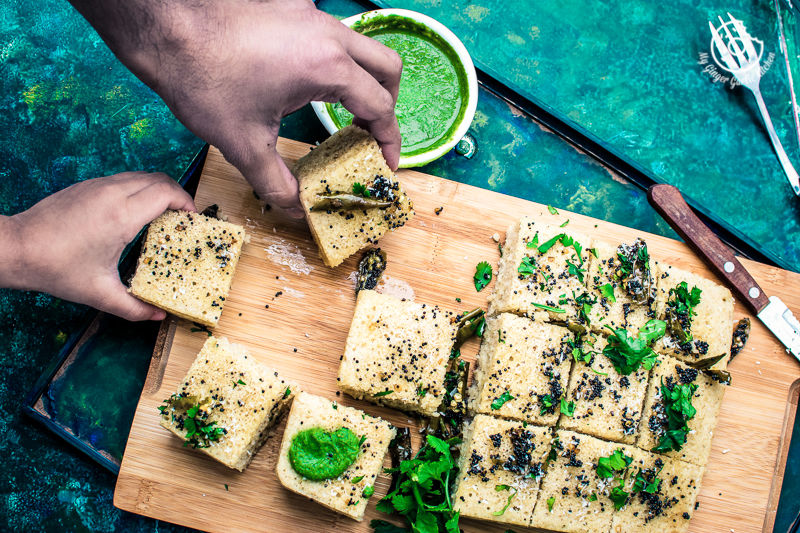 two hands taking two pieces of instant oats rava dhokla from a wooden board