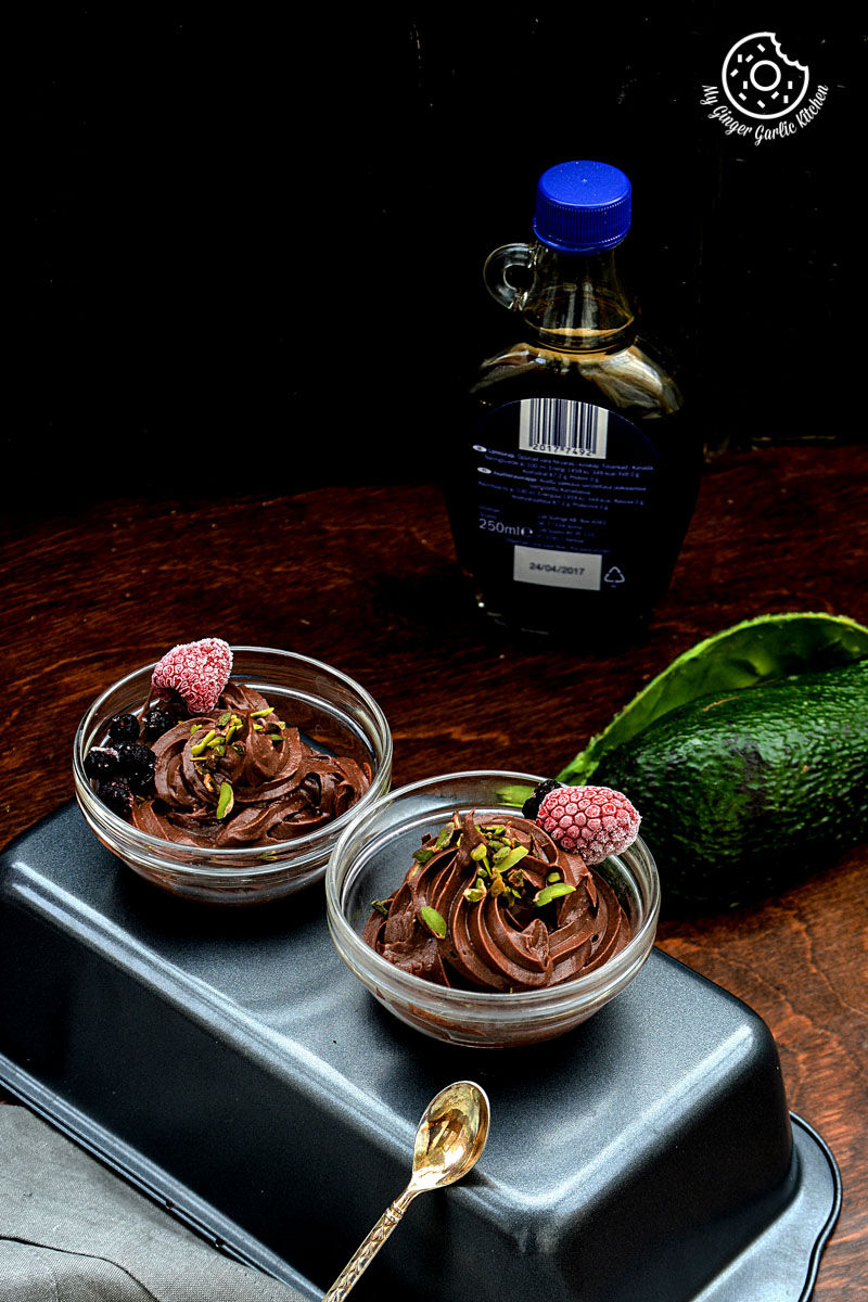 three bowls of dairy free avocado chocolate mousse with a spoon and a jar of syrup