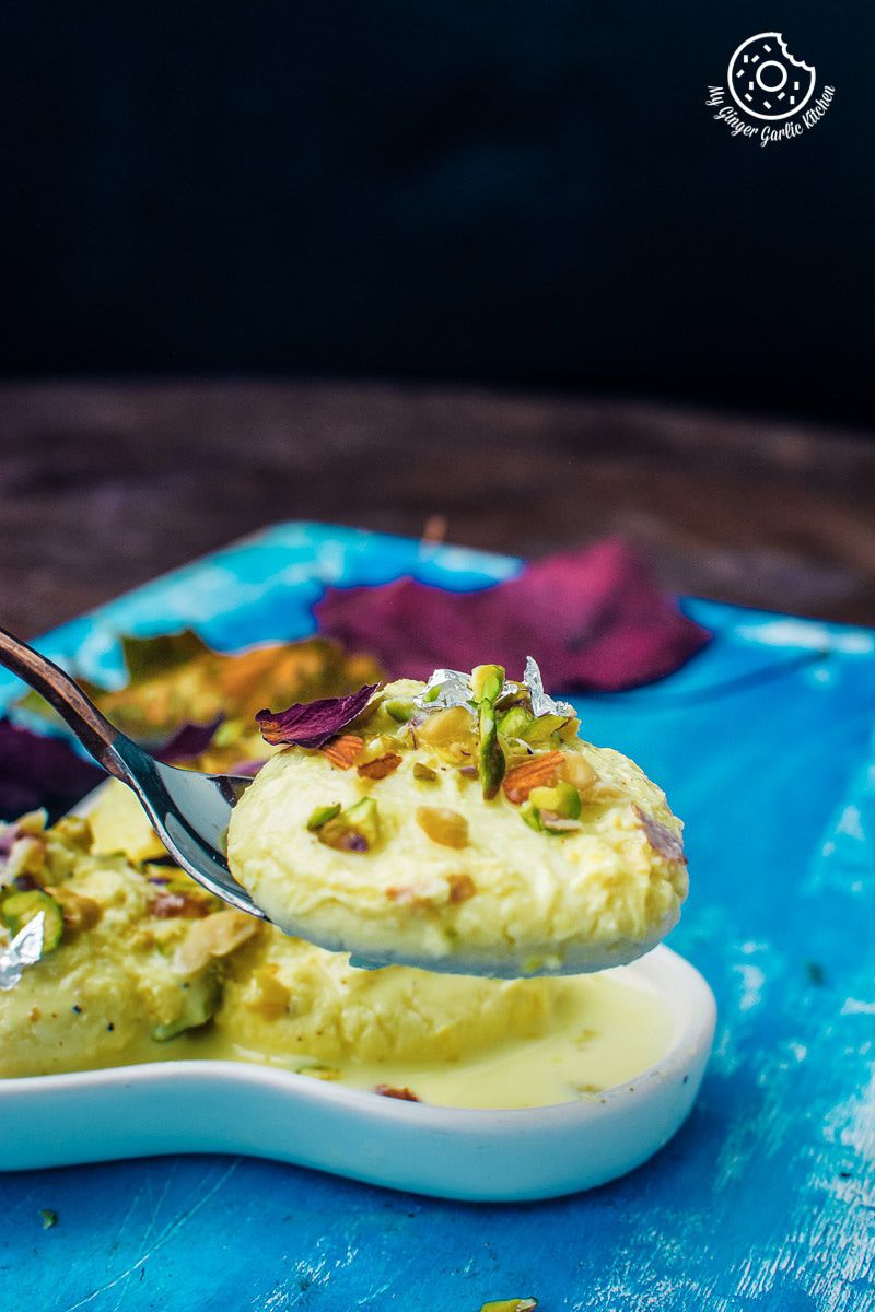 a spoon with ras malai on with a plate of soft rasmalai on a blue table