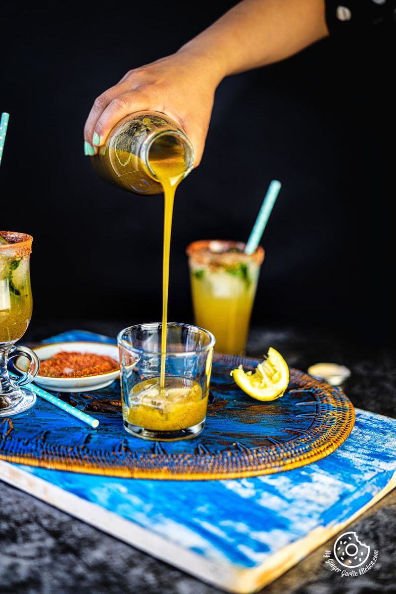 a hand pouring aam panna concentrate from a glass bottle into a transparent glass