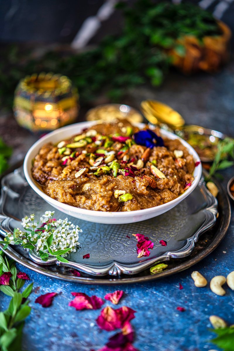 Rich Atta Halwa in a white ceramic dish, garnished with a colorful mix of nuts and rose petals, set against a rustic blue backdrop.