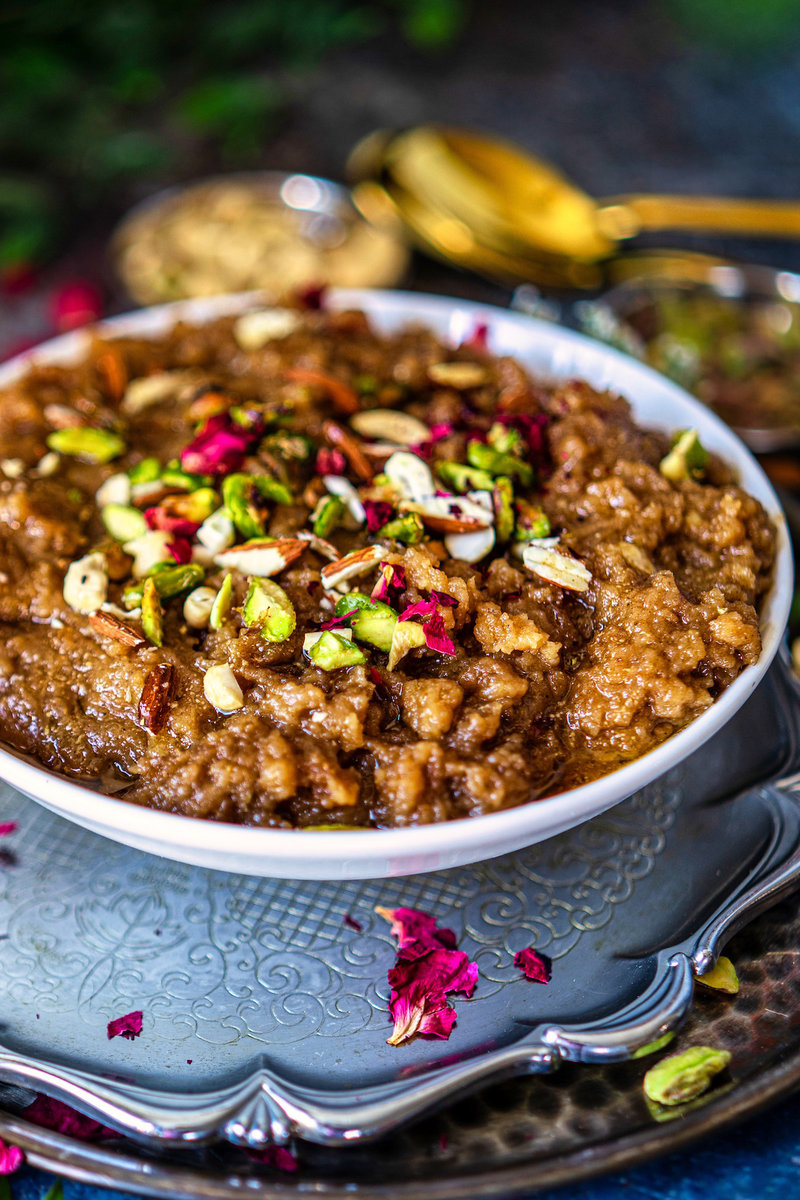 Aate Ka Halwa topped with chopped nuts and rose petals, served in a decorative white bowl on a patterned silver tray, surrounded by greenery and candlelight.