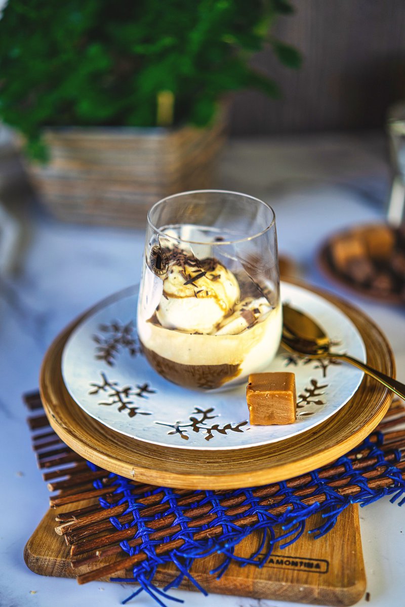 A glass of affogato dessert with vanilla ice cream and hot espresso, topped with dark chocolate shavings, served on a decorative plate with a caramel square on the side.