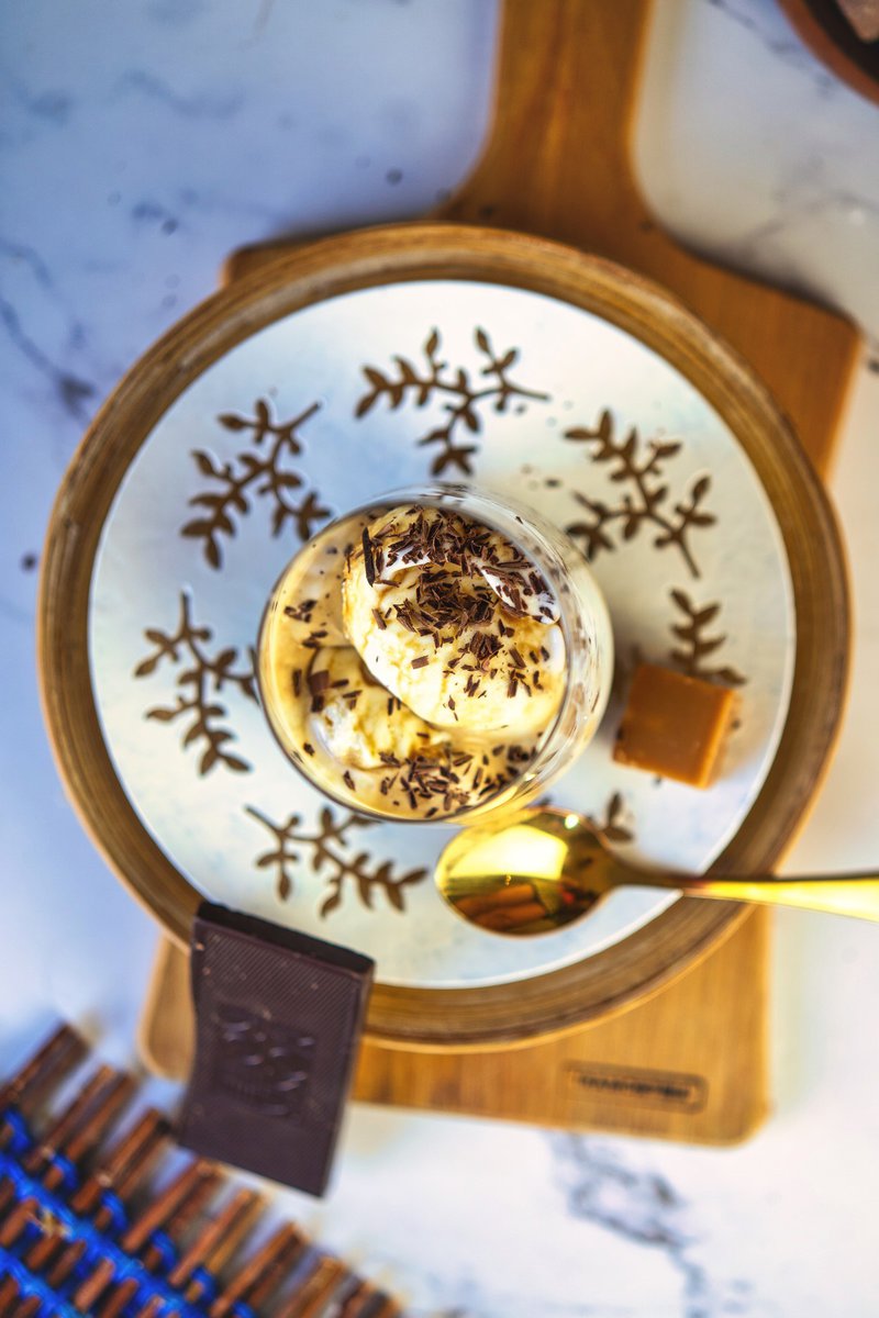 Top view of affogato served in a glass on a decorative plate, with vanilla ice cream, dark chocolate shavings, a gold spoon, and a piece of caramel on the side.