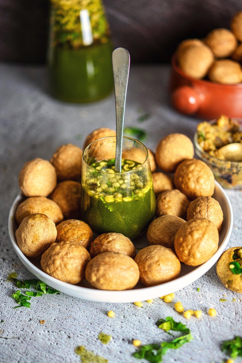 Plate of puris arranged around a glass filled with spicy green pani, with garnishes scattered around.