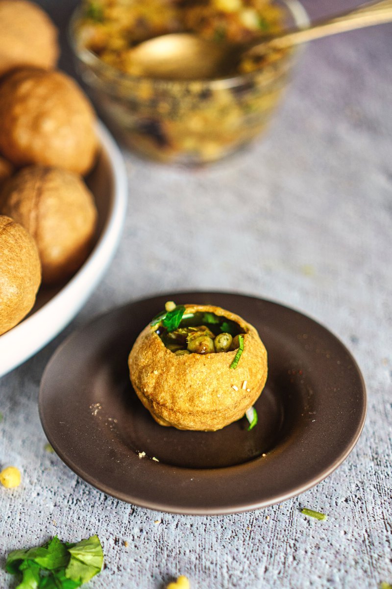 Close-up of a single puri filled with spicy filling on a small plate, with more puris and ingredients in the background.