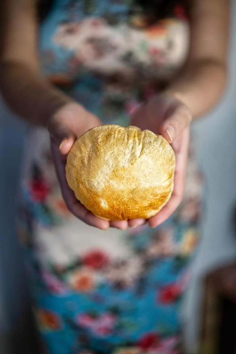 A person holding a perfectly puffed air-fried poori in their hands. The poori is round, fluffy, and golden brown. The person is wearing a blue dress with a floral pattern.