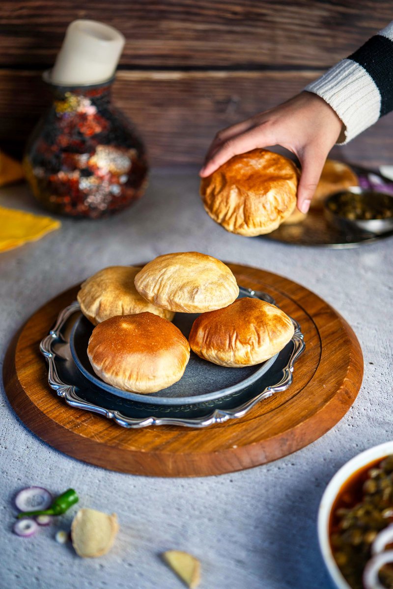 A hand reaching for a fluffy air-fried poori from a collection on a silver platter, with Indian dishes in the backdrop.
