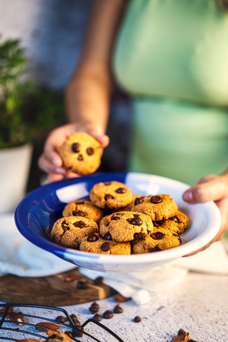 Soft and chewy almond flour tahini chocolate chip cookie held close to the camera, with more cookies in a bowl in the background.