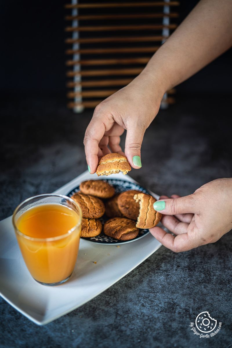 two hands holding two halve pieces of almond flour cookies with cookie plate and a juice glass