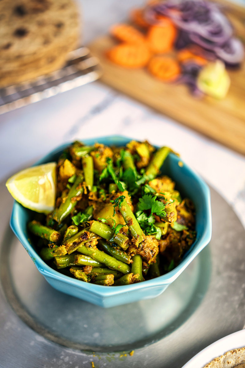 A bowl of Aloo Beans garnished with cilantro and a lemon wedge, with sliced vegetables and flatbreads in the background.