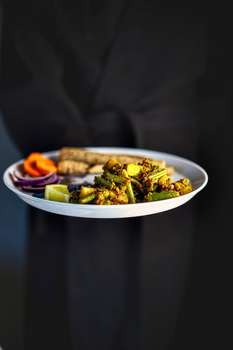 A person holding a plate of aloo beans garnished with vegetables, with a dark background.