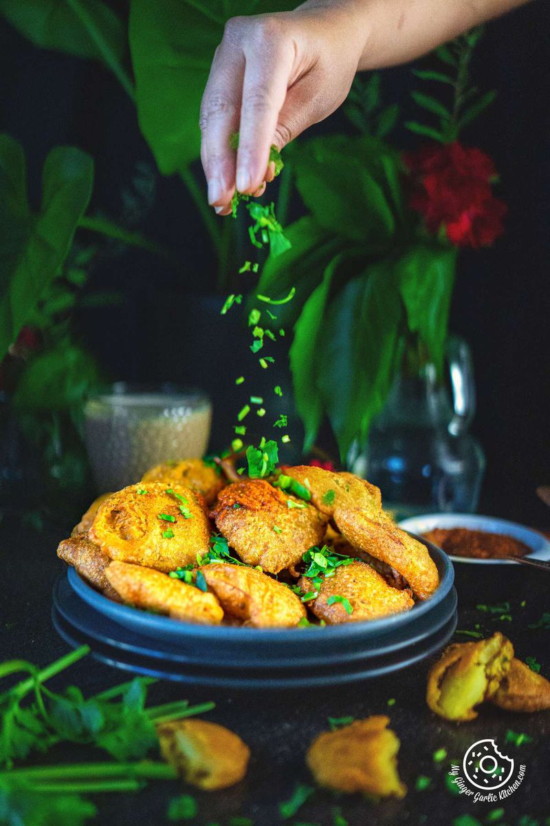 photo of a plate of fried aloo pakoras with a hand sprinkling them with green cilantro herbs