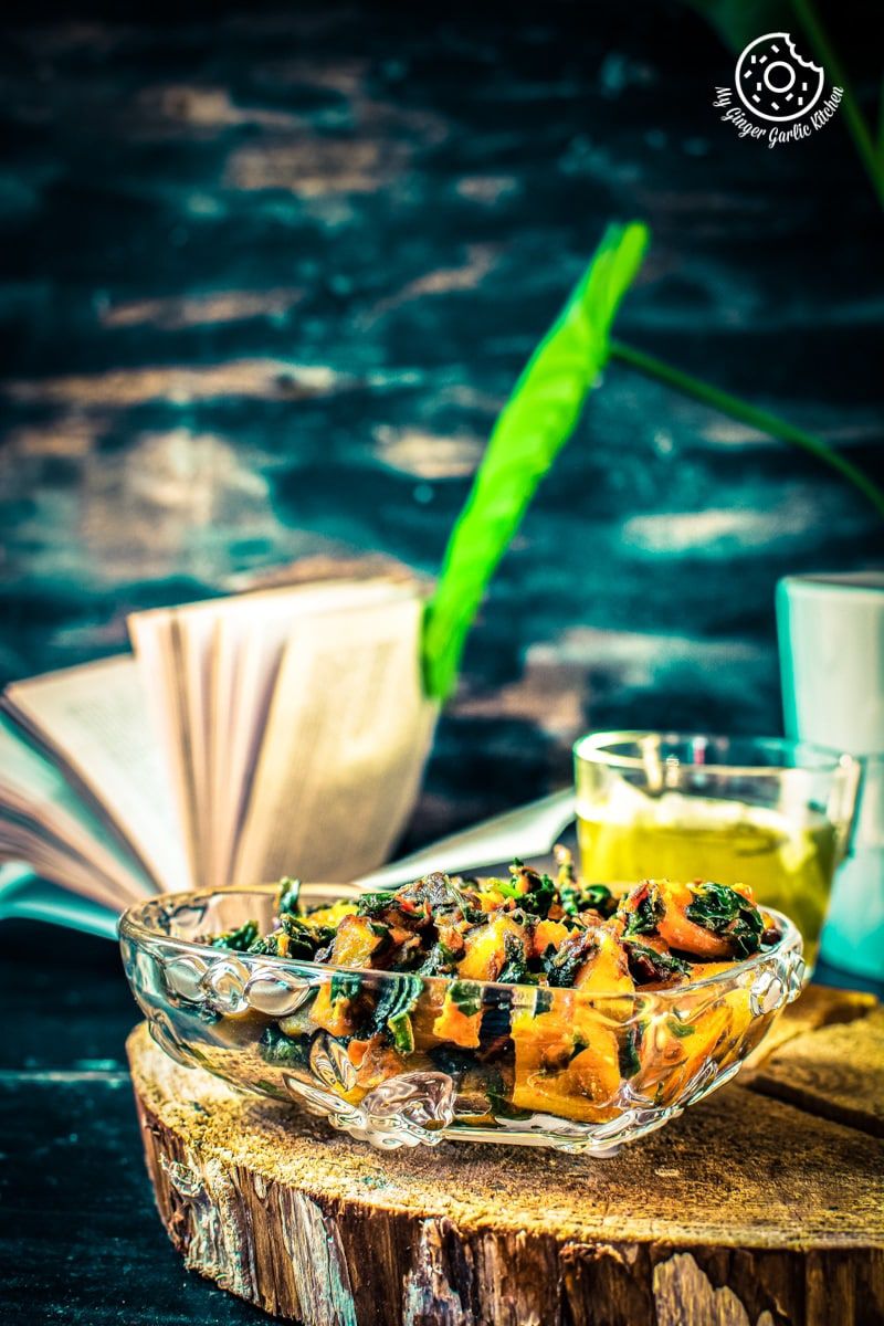 a glass bowl of aloo palak kept on a wooden log and a book in backdrop