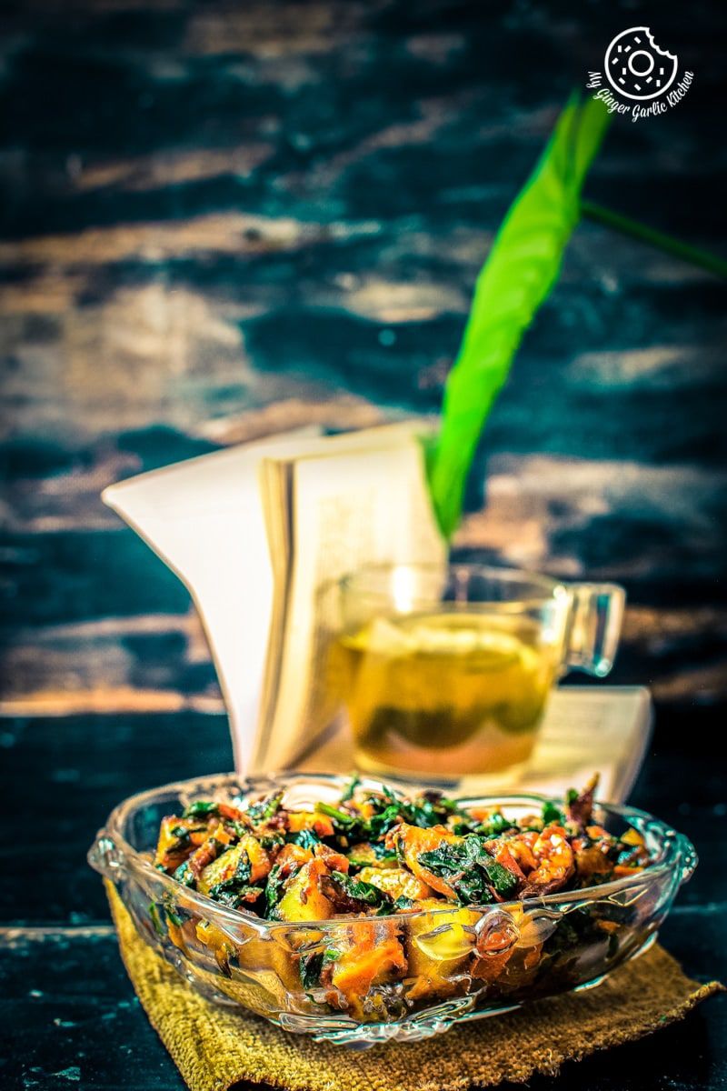 a glass bowl of aloo palak kept on a brown wooden log and a transparent tea cup in background