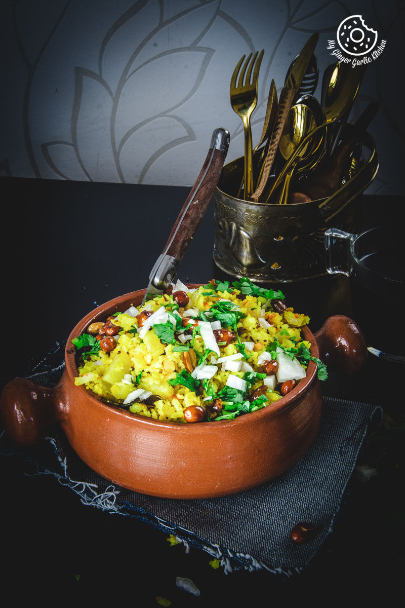 a bowl of aloo poha topped with peanuts, onions and lime wedge, and cutlery on a table