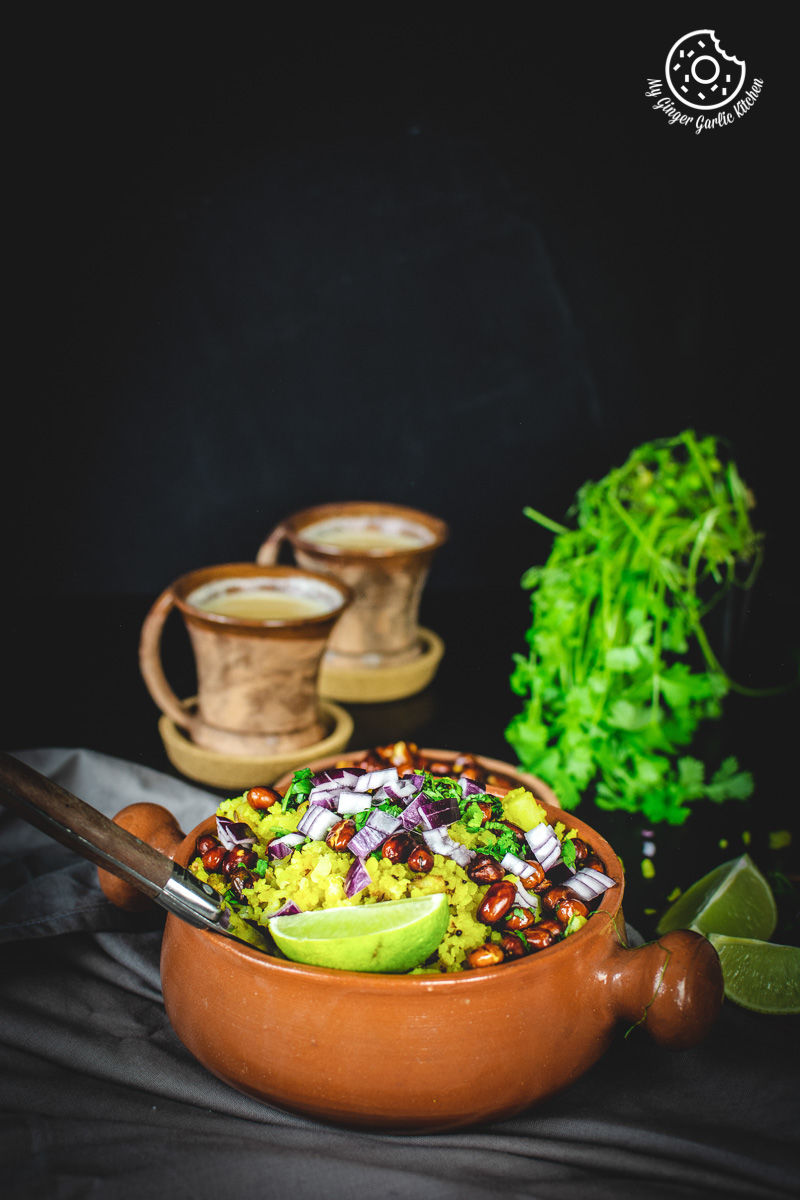 a bowl of aloo poha topped with peanuts, onions and lime wedge with a two cups of coffee in background