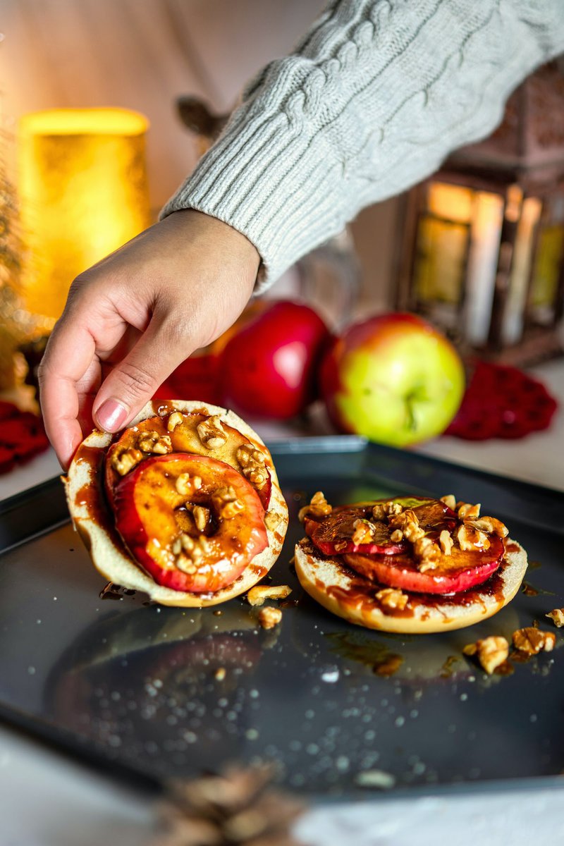 A hand placing a grilled apple slice with walnuts and syrup on a bagel, with a cozy, festive backdrop.
