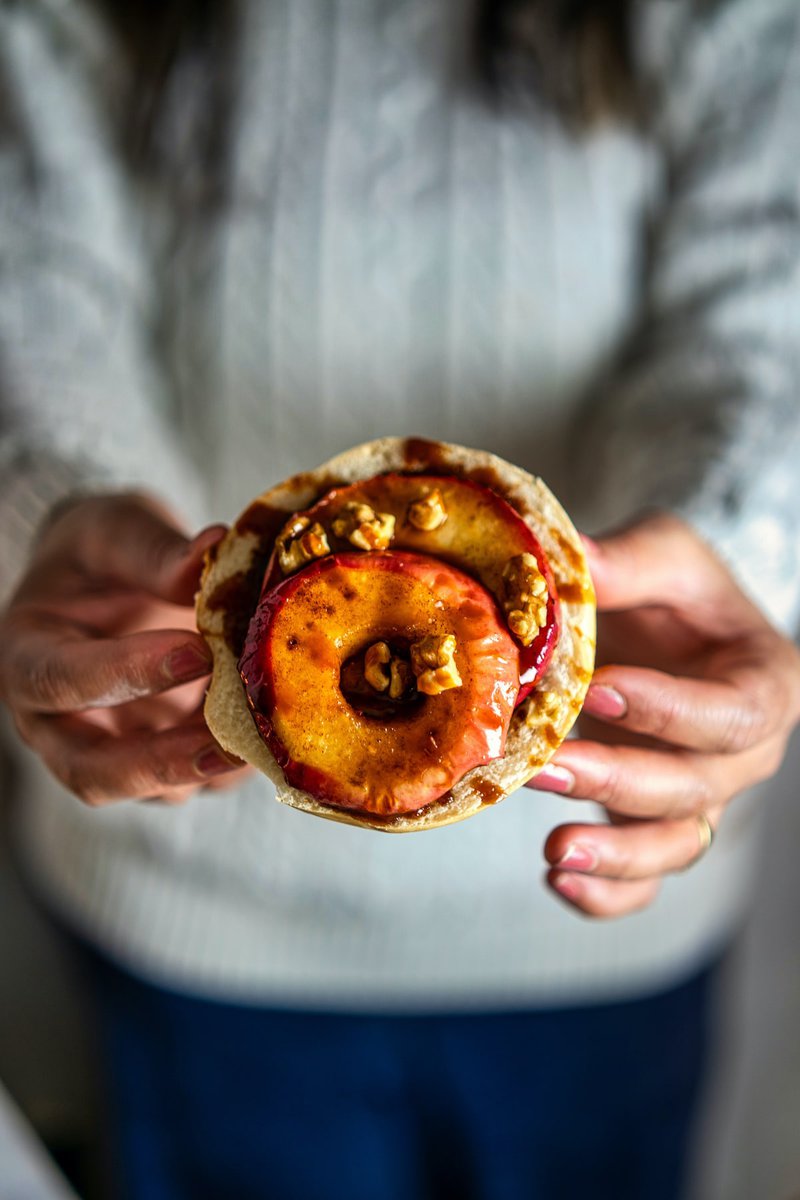 A person holding an open-faced apple bagel topped with a grilled apple slice, walnuts, and a drizzle of syrup.