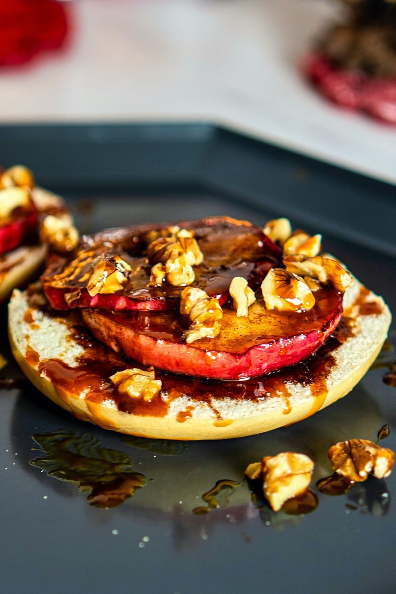 A close-up of a grilled apple bagel with walnuts and syrup on a black plate, with a soft-focused background.