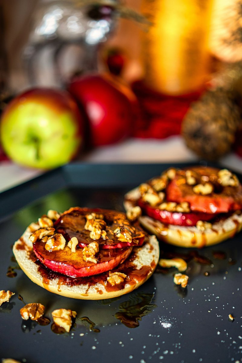 An open-faced bagel with a grilled apple slice and walnuts, drizzled with syrup, set against a festive background.