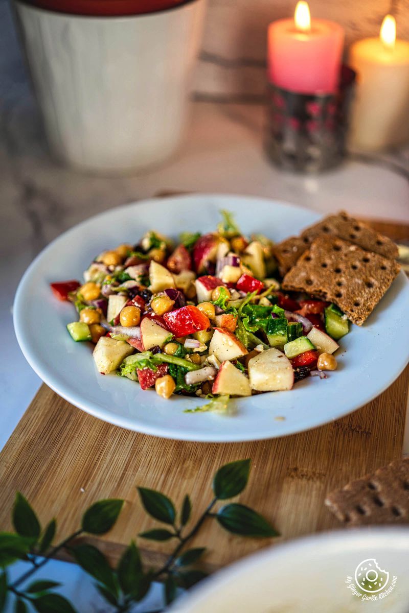 a white plate with a apple salad and cracker on it next to a candle in the background