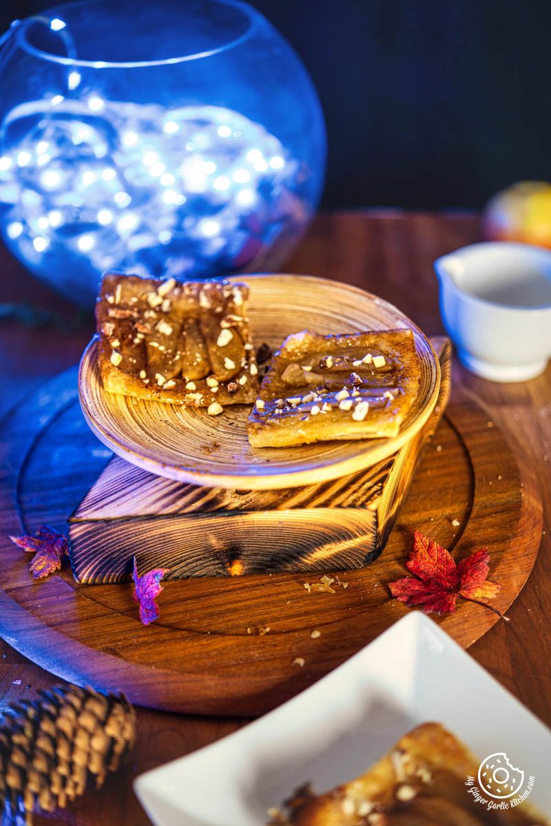 a wooden plate of apple upside down puff pastry sits on top of a wooden table with a bowl of lights in the background and some leaves in the foreground