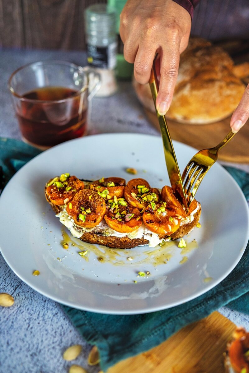 Grilled apricot toast with whipped cottage cheese, pistachios, and honey being sliced on a plate with golden fork and knife
