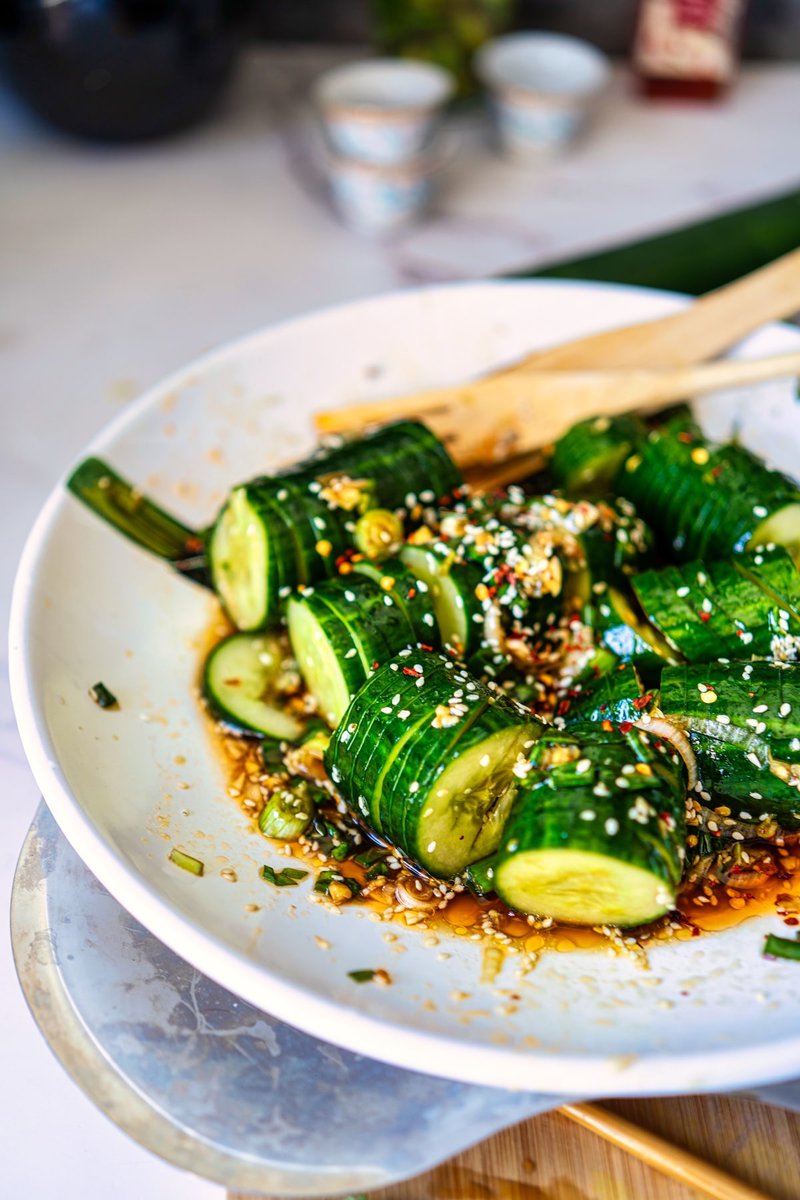 Close-up of Asian cucumber salad with sesame seeds and a tangy dressing in a white bowl, showing vibrant green cucumber slices and wooden utensils.