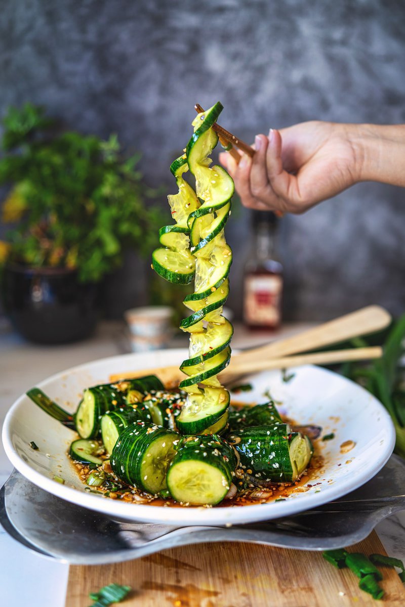 Hand holding a spiralized cucumber slice above a bowl of Asian cucumber salad, highlighting the fresh, crunchy texture and garnished with sesame seeds and a flavorful dressing.