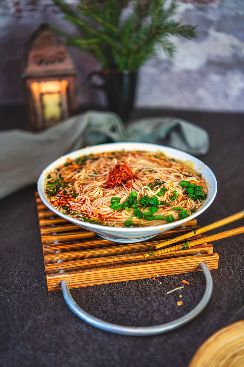  A bowl of Asian noodle soup on a wooden stand, garnished with chili flakes and green onions, with a dark, moody background.