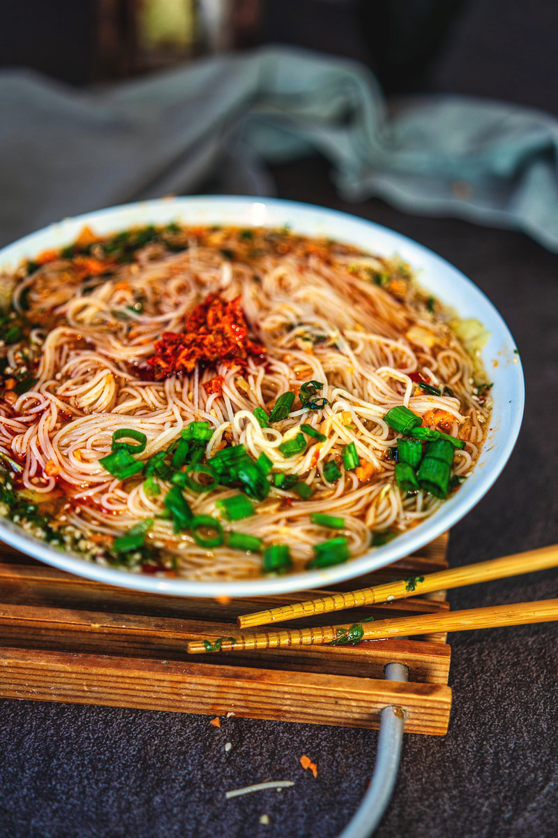 A bowl of Asian noodle soup with a rich broth, garnished with green onions and chili flakes, placed on a wooden stand with chopsticks on the side.
