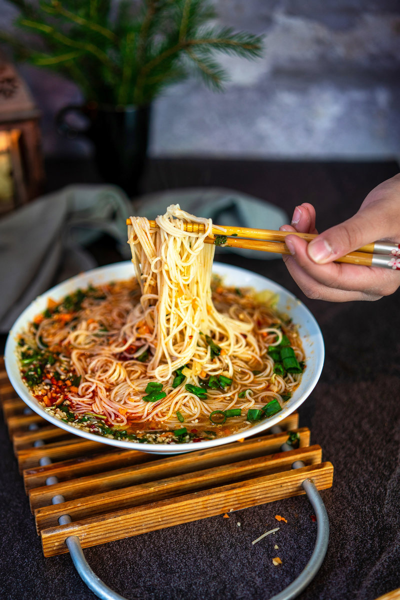 Someone using chopsticks to lift noodles from a bowl of Asian noodle soup, which is richly garnished with herbs and spices, on a wooden stand.