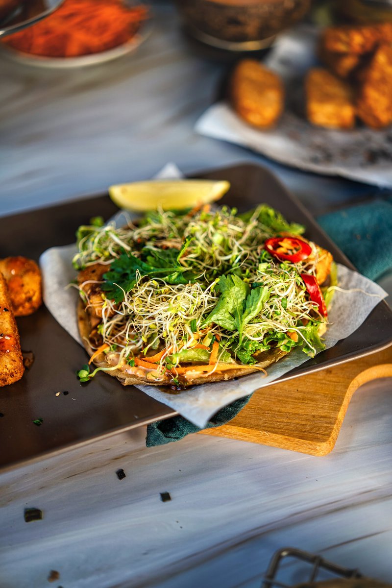 A vibrant Asian taco with fresh greens, sprouts, and chili slices on a dark plate.