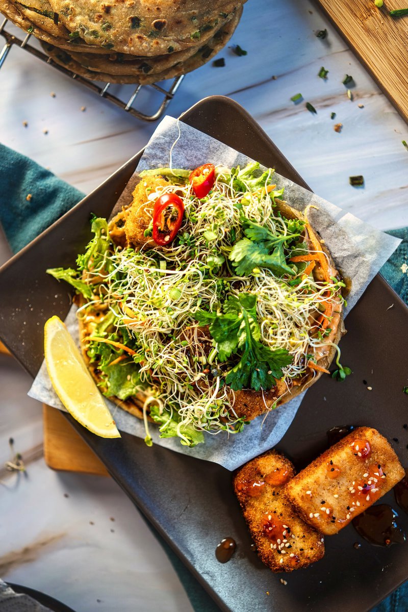 Asian taco with fresh greens, sprouts, and red chili slices, served with a lemon wedge and tofu on a black plate.