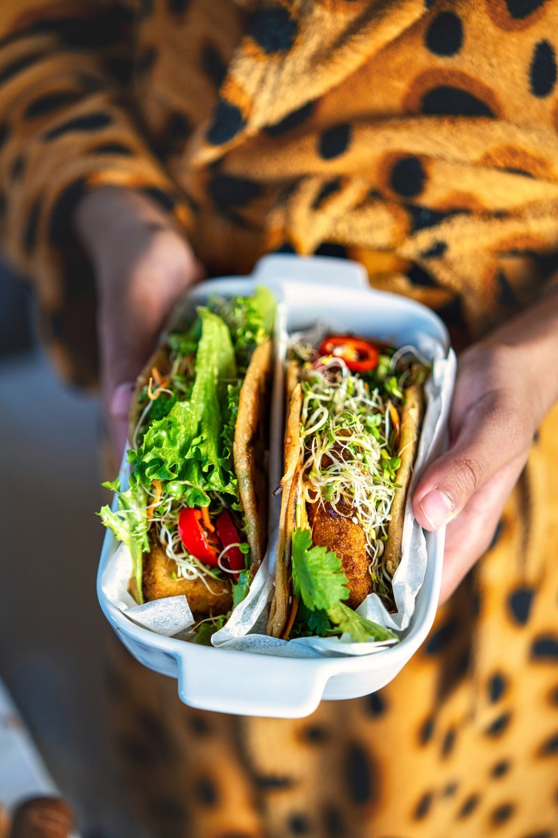 Close-up of hands holding a dish with two Asian tacos filled with fresh vegetables and sprouts.