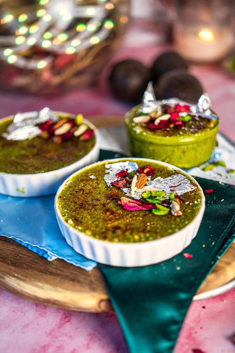 Creamy avocado burfi brûlée topped with silver leaf, rose petals, almonds, and pistachios in a white ramekin, held in hands