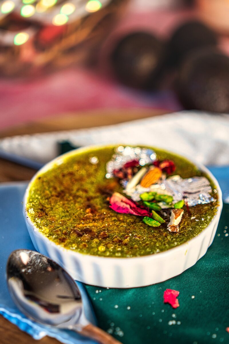 Close-up of avocado burfi brûlée with caramelized sugar crust, silver varq, rose petals, and sliced nuts served on a green satin cloth