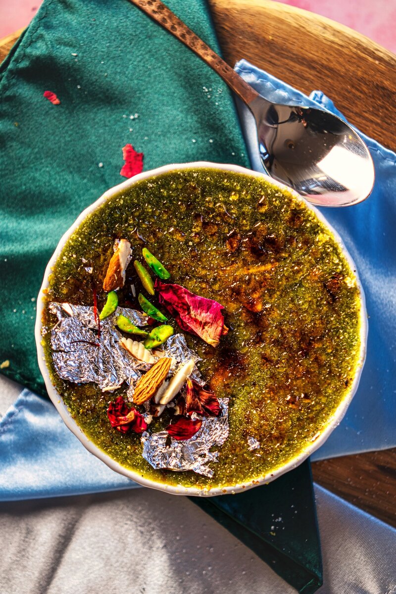 Avocado burfi brûlée dessert with silver leaf and nuts, served in white ramekins on satin fabric with spoon