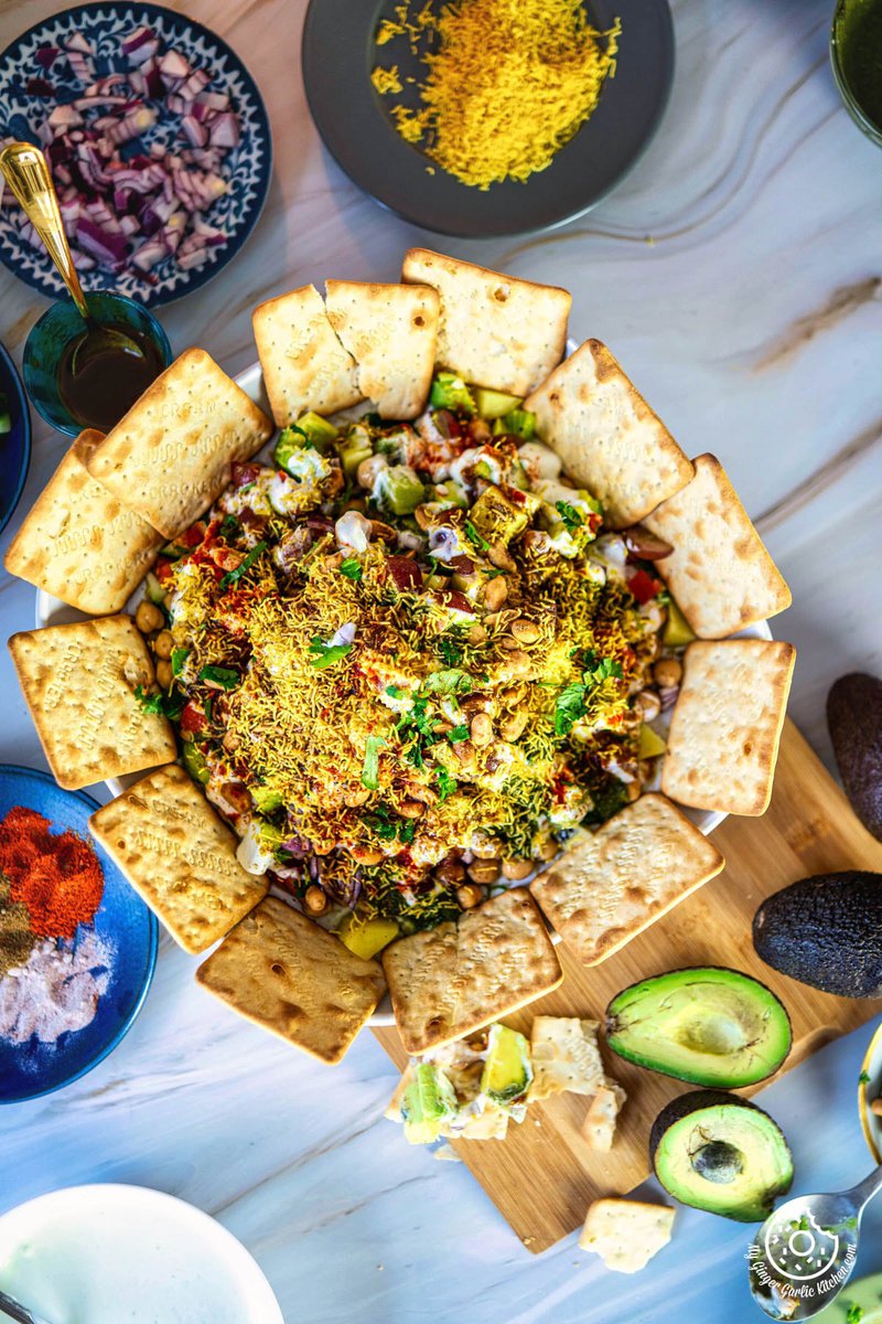 overhead photo of a plate of avocado chaat on a table with crackers, sliced avocados and other toppings on the side
