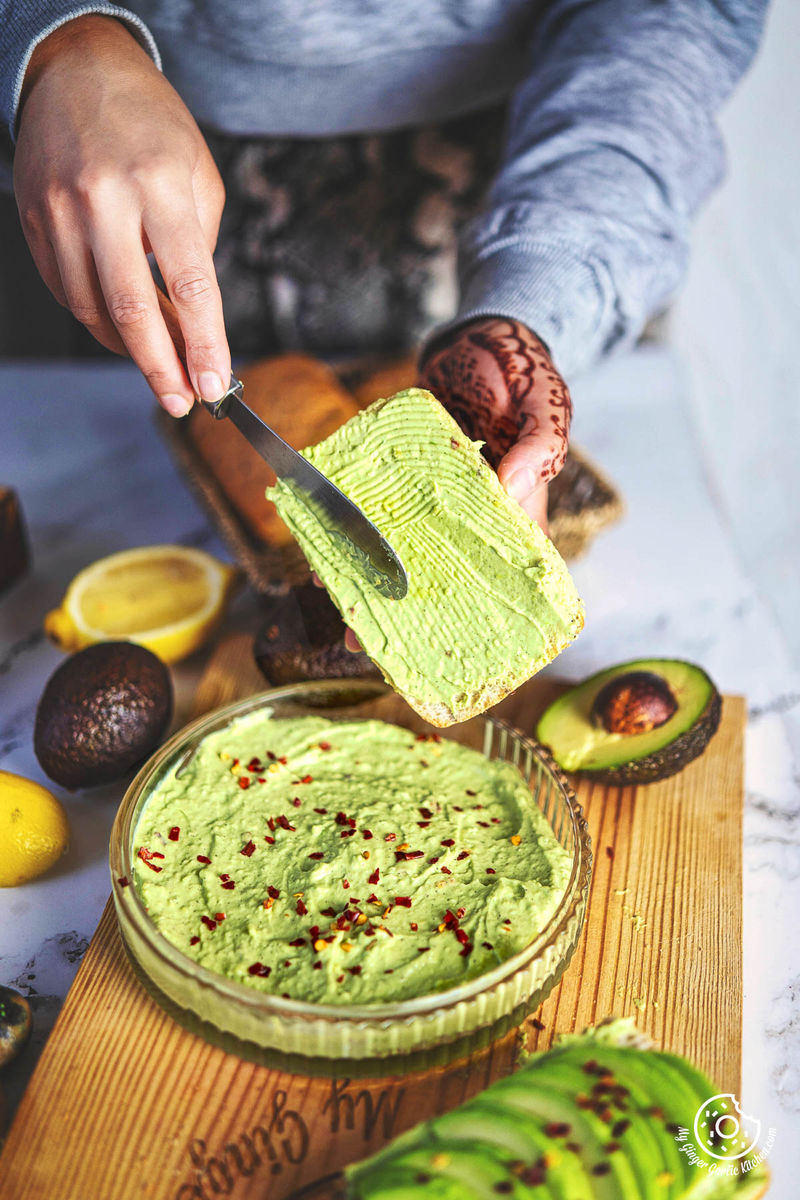 a person holding a bowl of avocado cheese spread in front of a table with other food and condiments