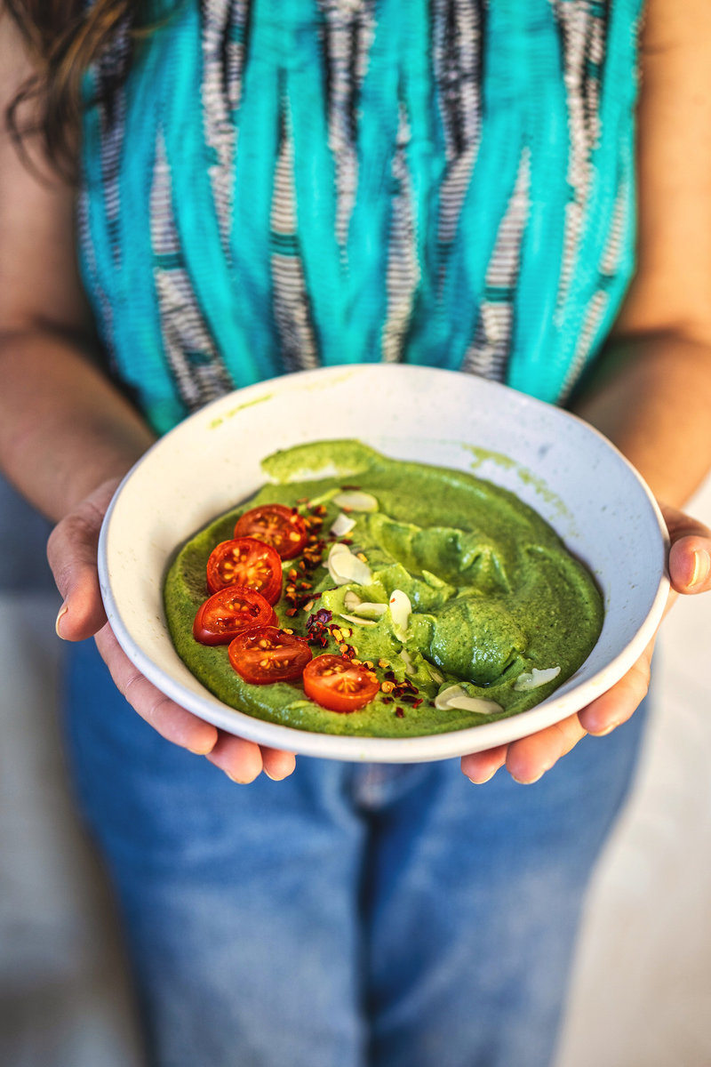 A person holding a bowl of avocado chutney garnished with cherry tomatoes, almond flakes, and chili flakes, ready to be served.