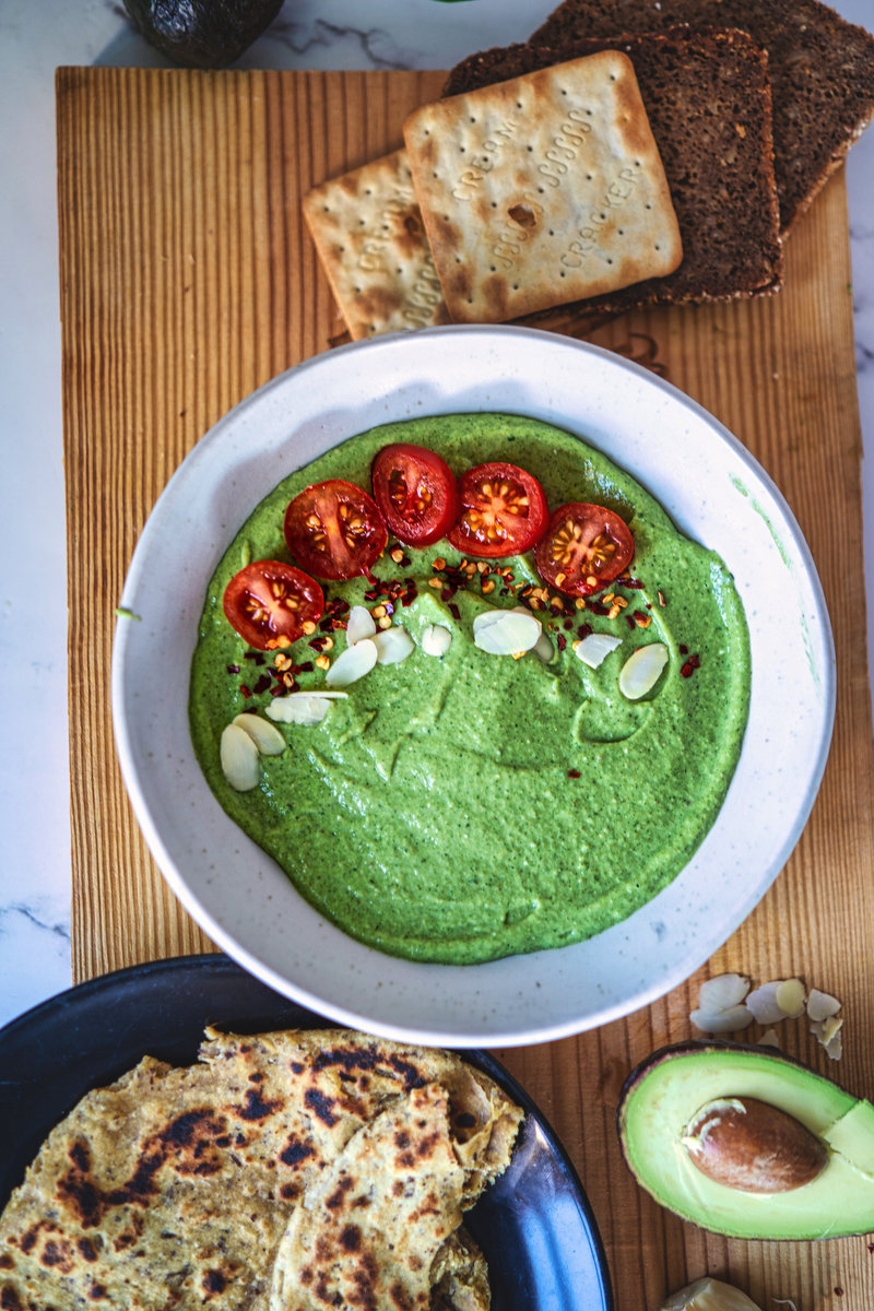 A bowl of creamy avocado chutney topped with sliced cherry tomatoes, almond flakes, and red pepper flakes, served with rye bread and crackers on a wooden mat.