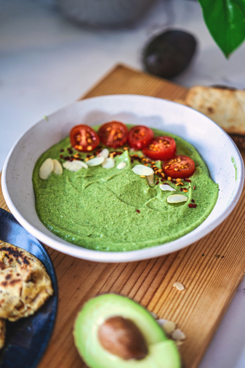 Close-up of a bowl of avocado chutney garnished with cherry tomatoes, almond flakes, and red pepper flakes on a wooden board with pita bread on the side.