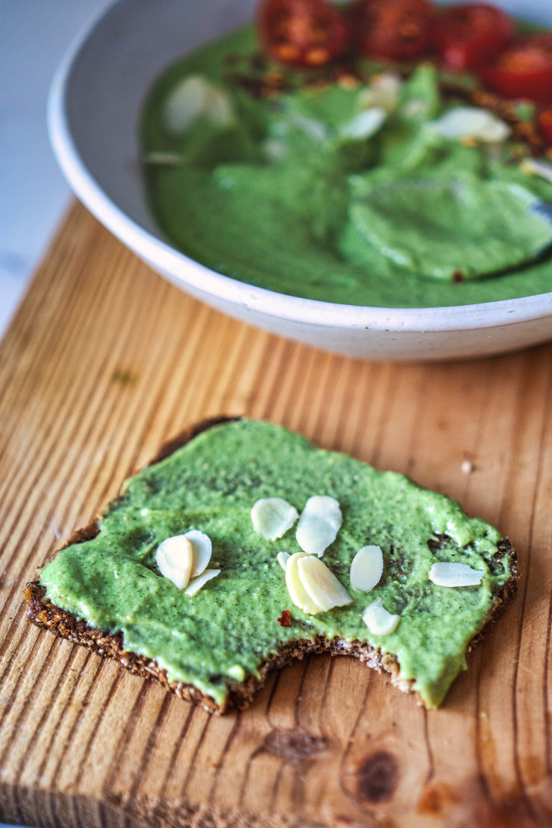 Avocado chutney spread on a piece of rye bread, topped with almond flakes, with a bowl of the chutney in the background on a wooden board.